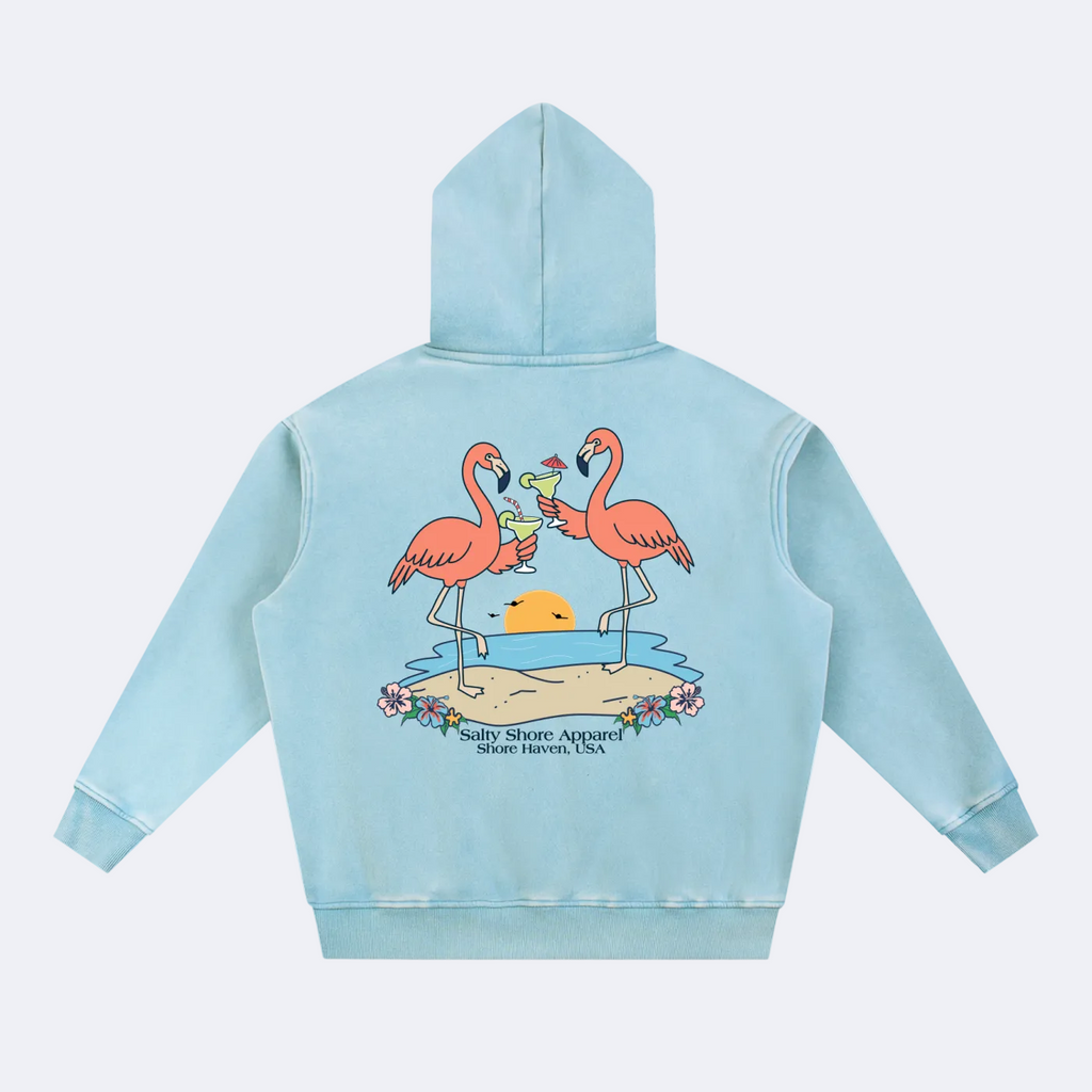 A light blue hooded sweatshirt shown from the back, featuring a graphic print of two pink flamingos standing on a beach holding margaritas with a yellow sun and the text 'Salty Shore Apparel Shore Haven, USA'.
