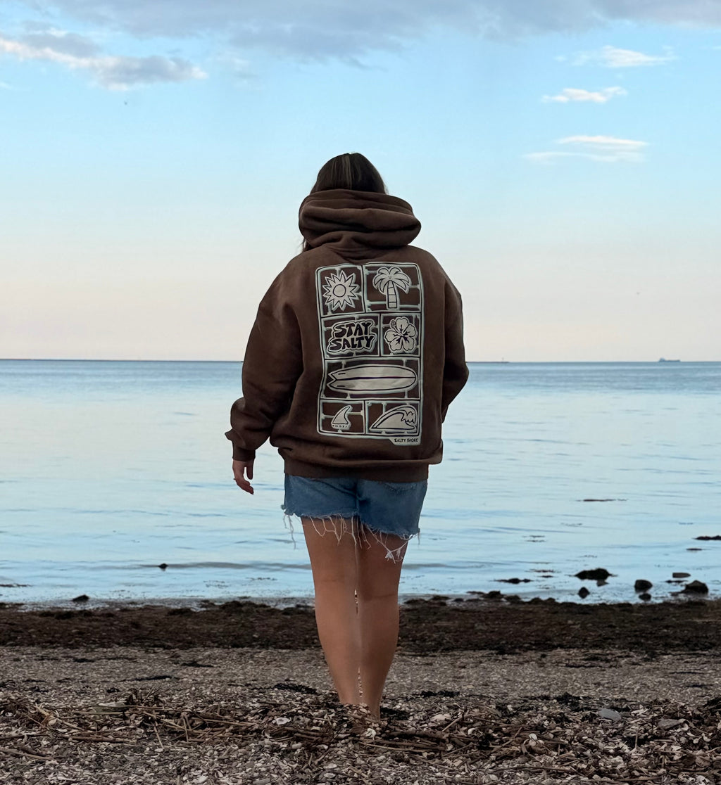 A person standing on a pebbled beach facing the ocean, wearing a brown hoodie with a large white graphic print on the back and denim shorts.