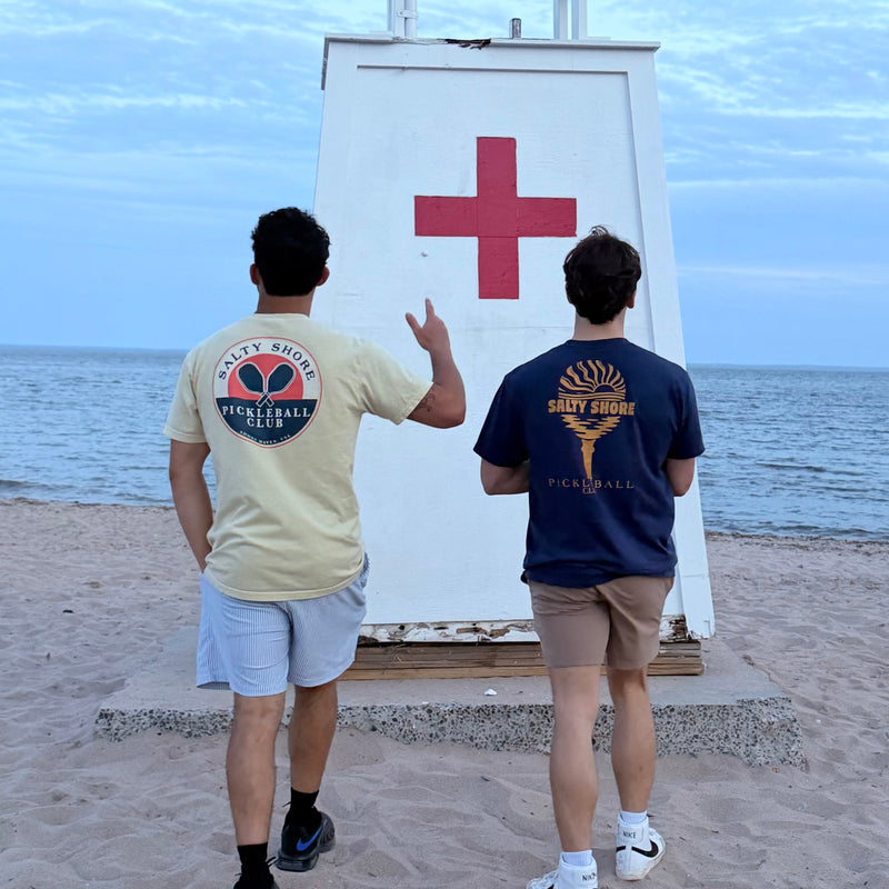 Two people on a beach pointing towards a white lifeguard stand with a red cross symbol.
