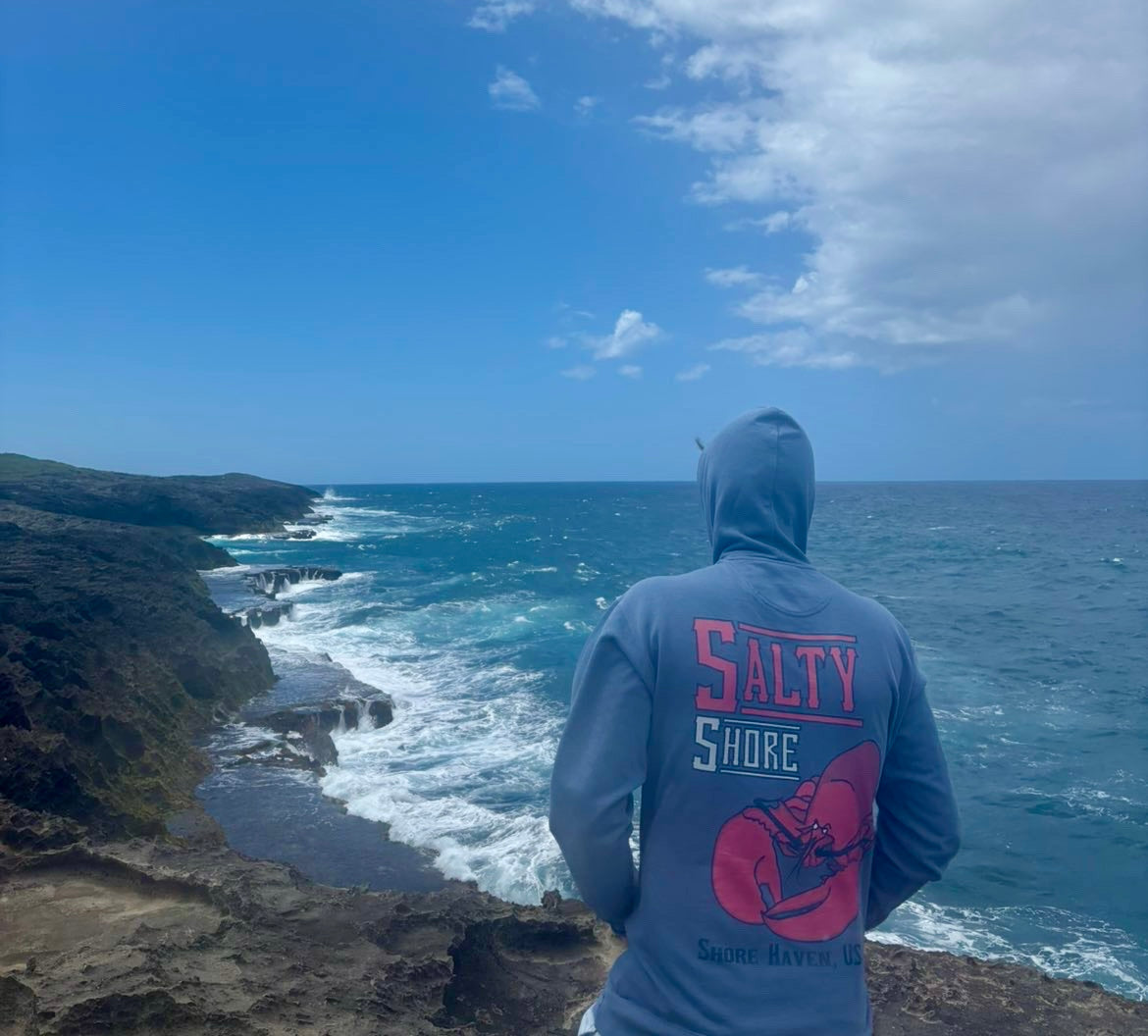 Person wearing a hoodie with 'Salty Shore' text and graphic, standing on a cliff overlooking the ocean.