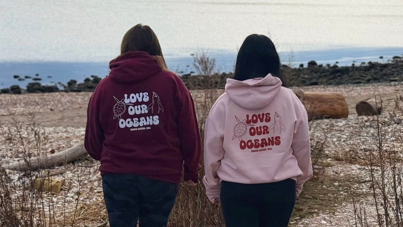 Two people wearing 'Love Our Oceans' hoodies walking along a pebbly path by the sea.