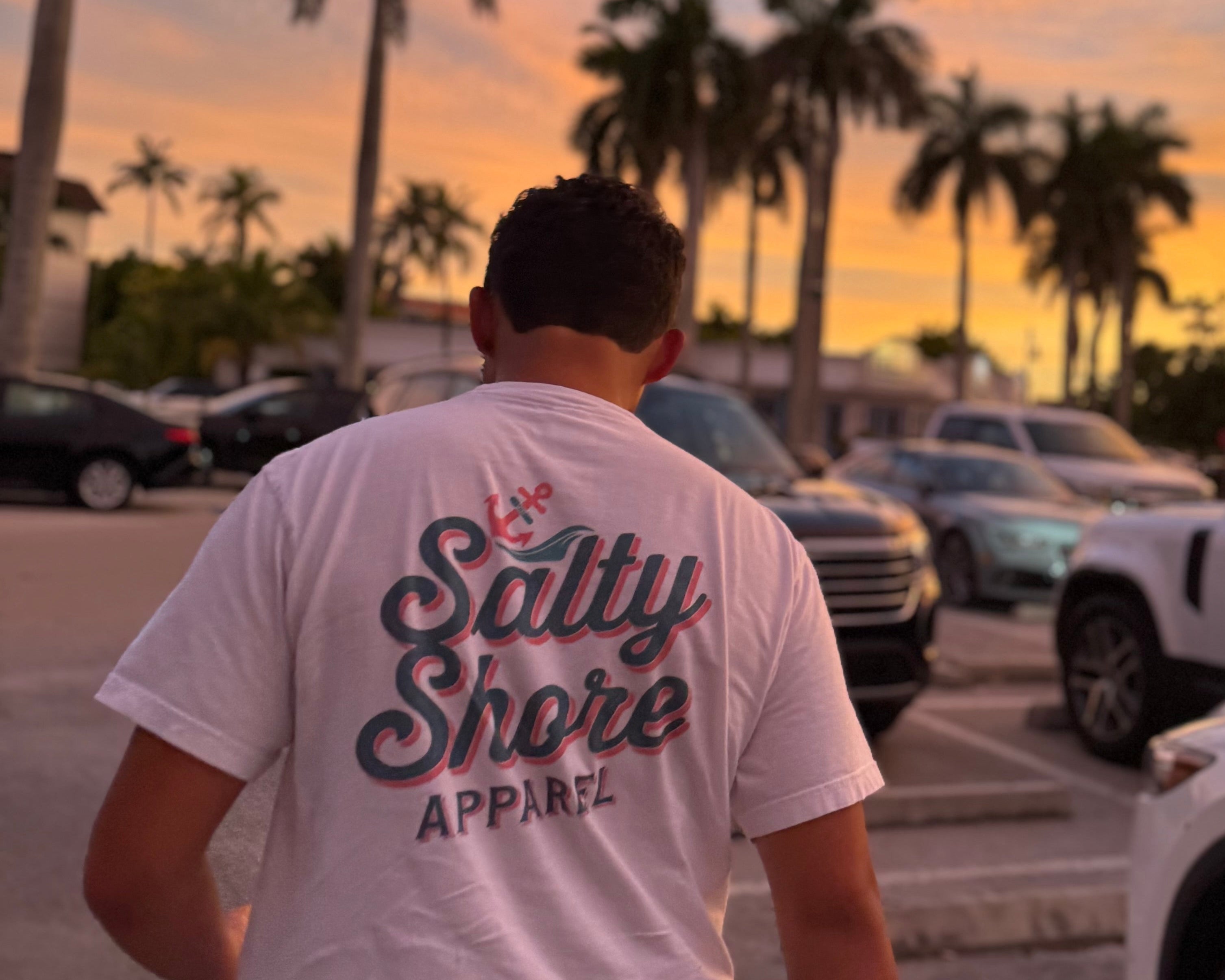 Person wearing a 'Salty Shore Apparel' t-shirt walking in a parking lot with palm trees and sunset in the background.