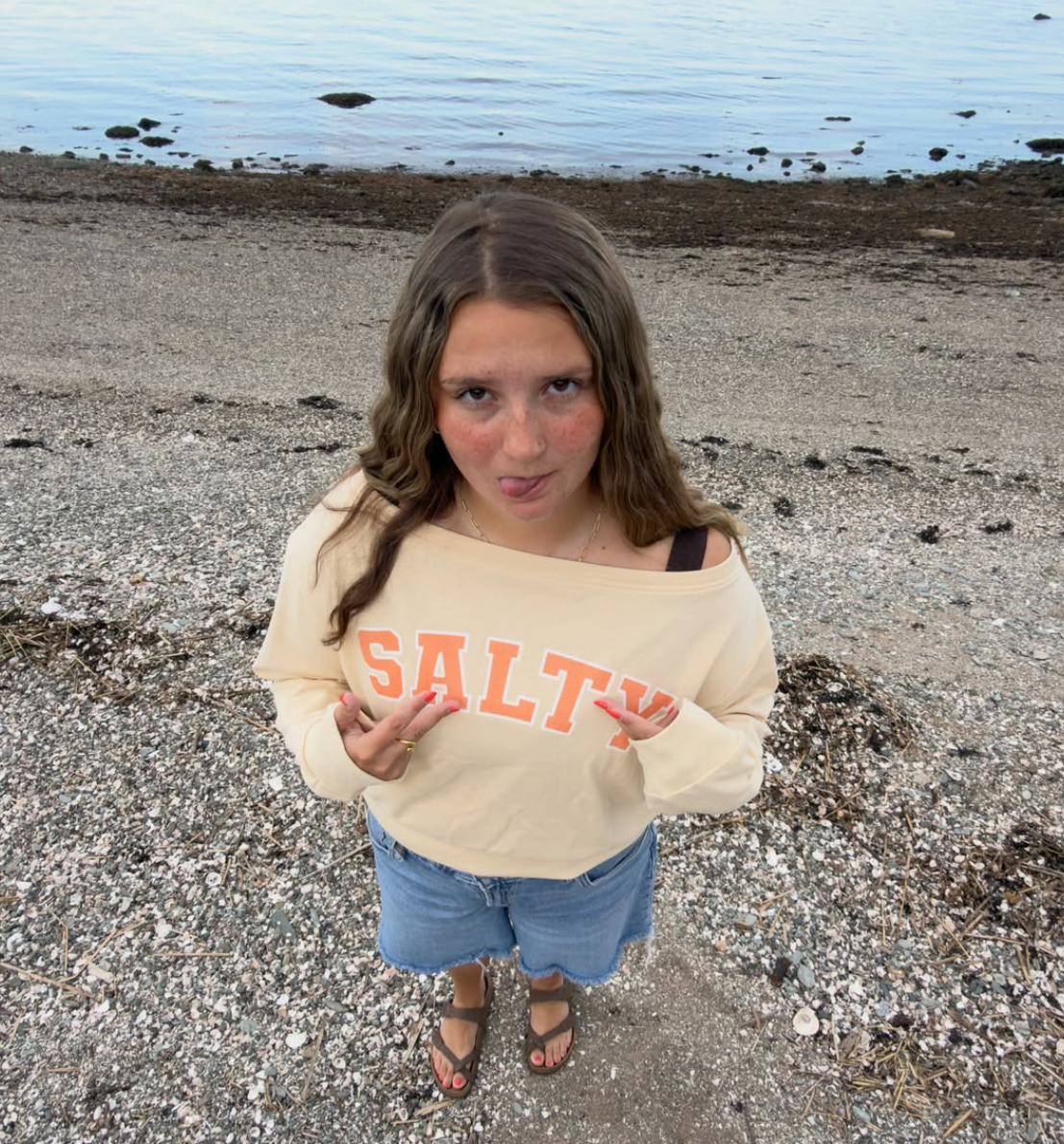 A screenshot of a Safari browser showing a Instagram post from saltyhomerapperd, featuring a young woman on a pebble beach wearing a cream off-the-shoulder sweatshirt with 'SALTY' printed in orange across the chest.